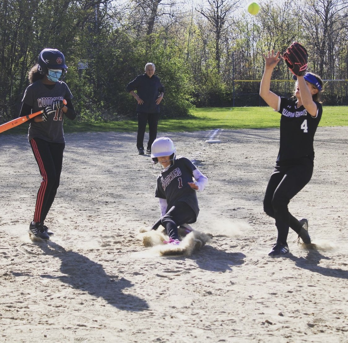 picture of a softball player sliding onto base while another player watches a third player catch the ball